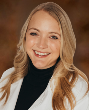 Clearwater Dental | Smiling woman with long blonde hair wearing a white coat and black turtleneck against a warm brown background